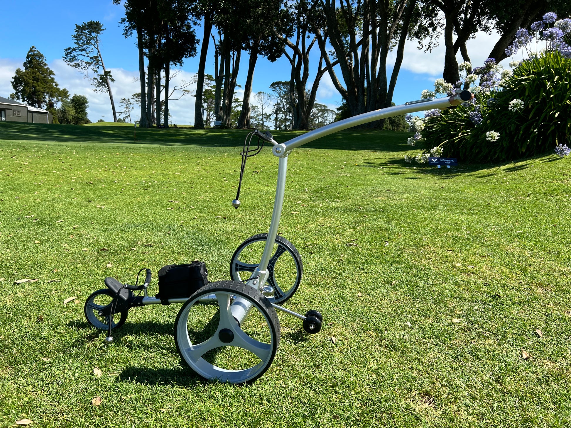 Golf push cart on a grassy field with trees and flowers in the background